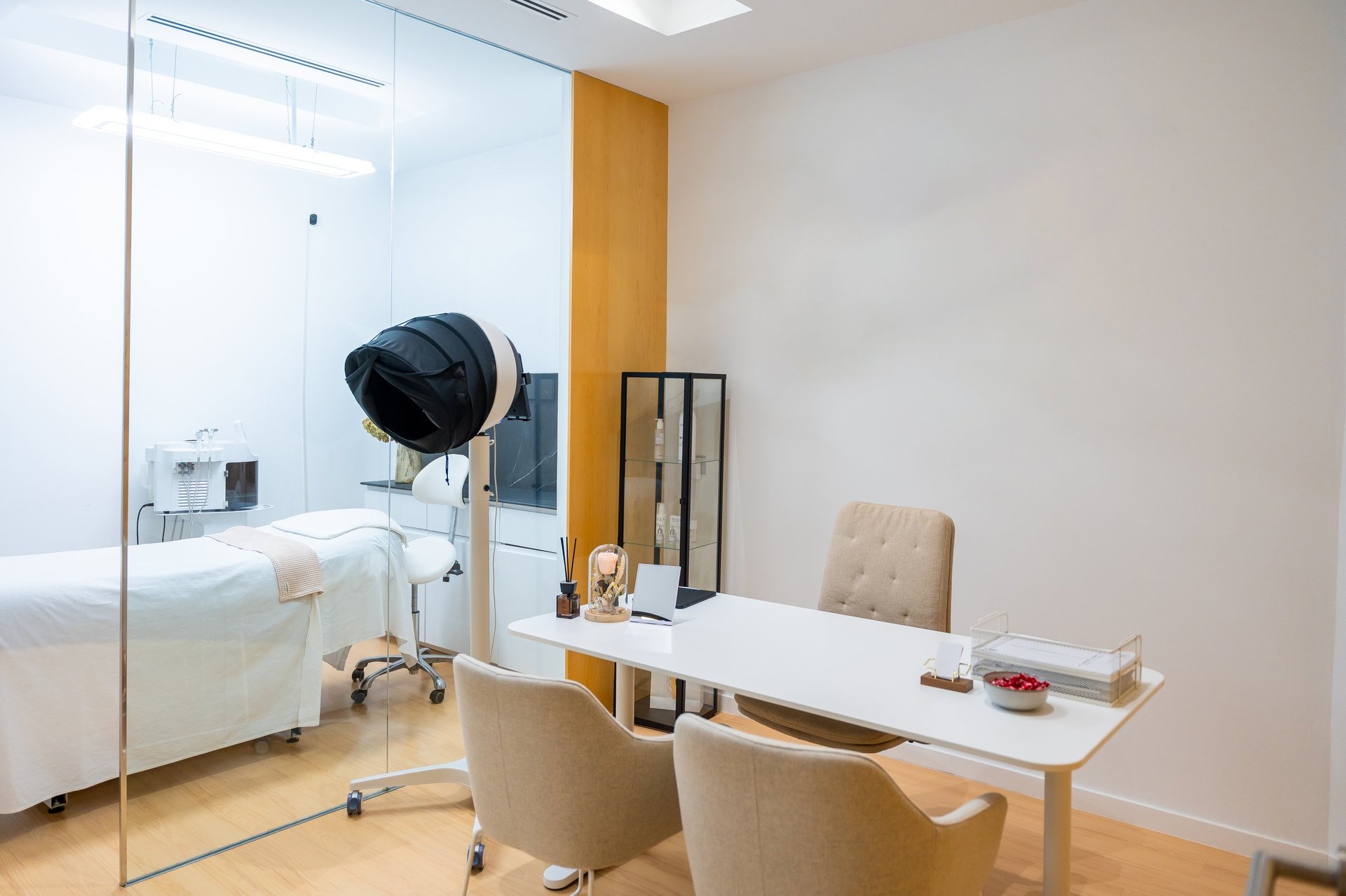 Contemporary aesthetic clinic featuring a consultation area with a white desk and two chairs, separated by a glass wall from a treatment room with a bed and specialized equipment